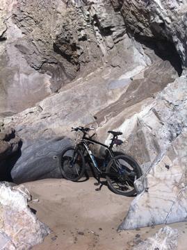 Specialized Hardrock Sport Disc: A black bicycle leaning against large, textured rocks on a sandy beach. The scene captures a rocky coastal area with natural formations and a hint of ocean in the background.