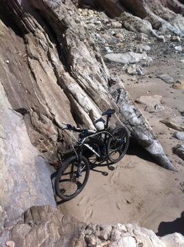 Specialized Hardrock Sport Disc: A black mountain bike leaning against a large rock formation on a sandy beach, surrounded by smooth stones and cliffs.