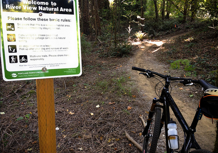 A mountain bike is parked next to a sign that welcomes visitors to the River View Natural Area. The sign lists basic rules for enjoying the area, including staying on trails and keeping dogs on leashes. The background features a winding dirt path through a lush green forest.