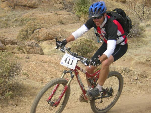 Iron Horse Hollowpoint Expert: A cyclist riding a mountain bike on a dirt trail in a rocky landscape. The cyclist, wearing a helmet and sport attire, is focused and smiling, while a race number is attached to the bike. Surrounding vegetation includes sparse shrubs and rocky outcroppings.