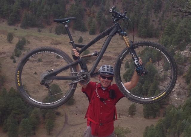 Yeti SB95: A cyclist wearing a helmet and a red shirt holds a mountain bike above his head while standing on a rocky outcrop, with a scenic view of forested hills in the background.