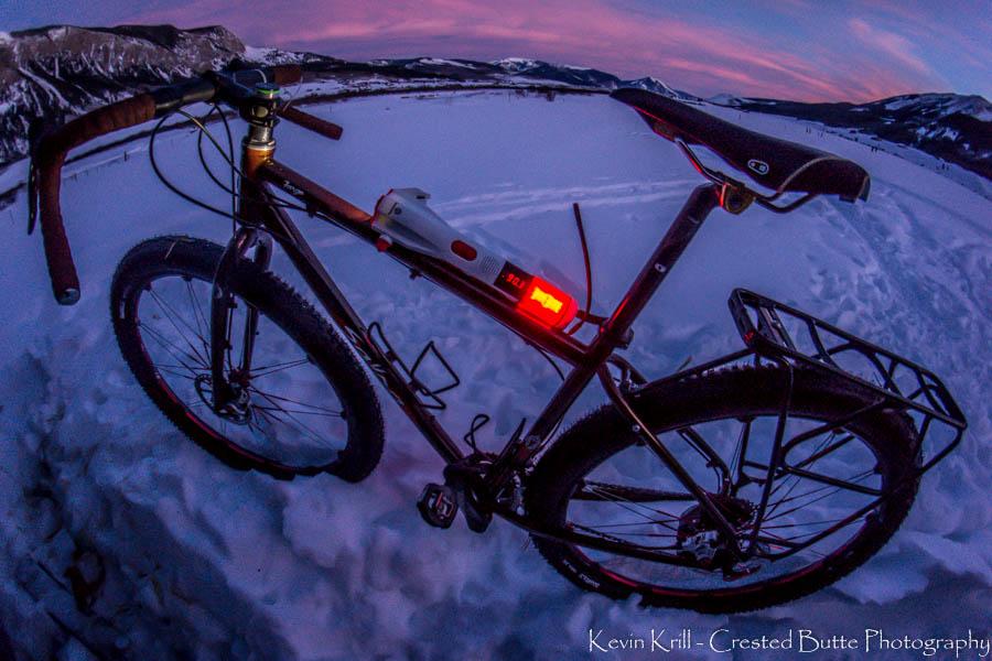 A fat tire bike stands on a snowy landscape at dusk, illuminated by a warm glow from its rear light. Snowy mountains are visible in the background, with the sky transitioning to twilight colors. The bike features a sturdy frame and handlebars, highlighting its readiness for winter terrain adventures.