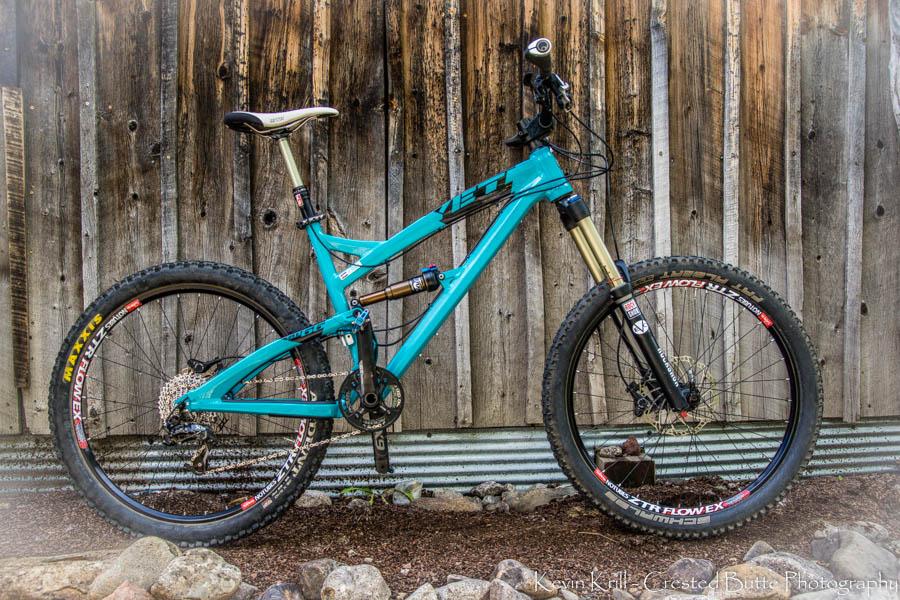 Yeti SB66: A turquoise mountain bike with a black frame, positioned against a rustic wooden fence. The bike features thick tires designed for off-road use, a gold suspension fork, and is equipped with various components for mountain biking.
