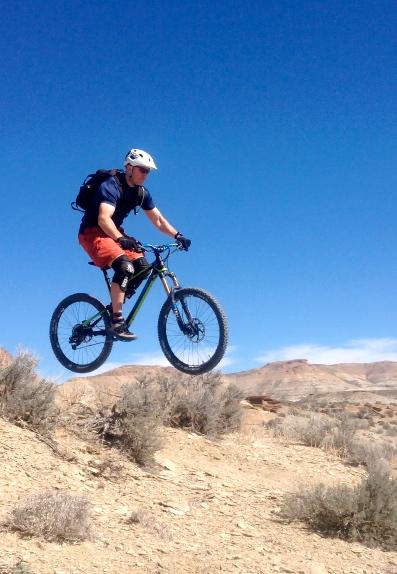 A person jumping on a mountain bike over a dirt trail, wearing a helmet and sporty clothing. The background features a clear blue sky and rocky terrain. Wilkins Peak Trails mountain bike trail.