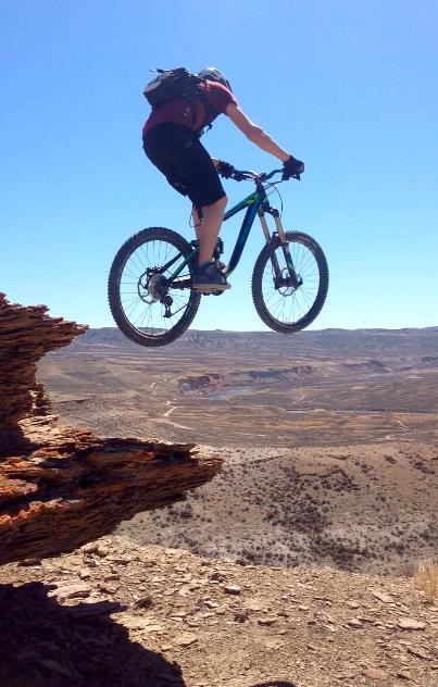 A mountain biker leaping off a rocky ledge into a desert landscape, showcasing a sense of adventure and skill. The background features rolling hills and distant terrain under a clear blue sky. Wilkins Peak Trails mountain bike trail.