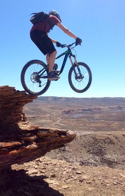 A mountain biker in mid-air, jumping off a rocky outcrop overlooking a vast, arid landscape. The rider is wearing a helmet and riding gear, with a clear blue sky above and distant hills in the background. Wilkins Peak Trails mountain bike trail.
