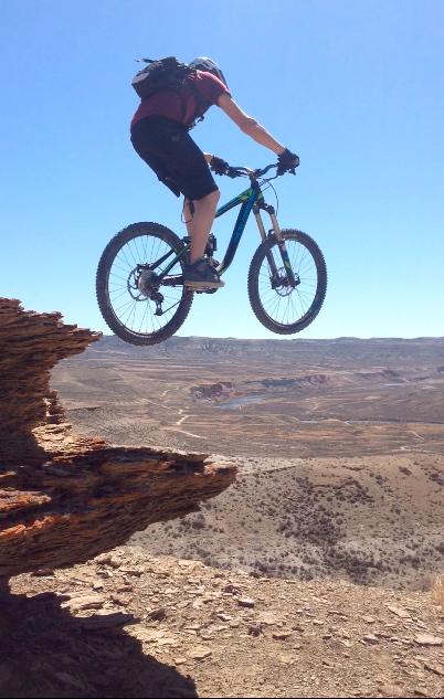 A mountain biker in mid-air performing a jump off a rocky ledge, with a vast desert landscape visible in the background under a clear blue sky. Wilkins Peak Trails mountain bike trail.