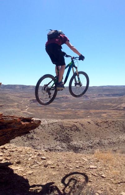 A mountain biker mid-air, performing a jump over a rocky edge with a vast, arid landscape in the background. The sky is clear and blue, highlighting the rugged terrain below. Wilkins Peak Trails mountain bike trail.