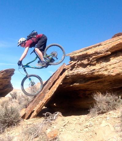 A mountain biker performing a jump off a rocky ledge in a rugged desert landscape under a clear blue sky. Wilkins Peak Trails mountain bike trail.