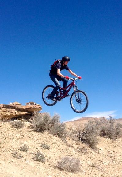 A mountain biker wearing a helmet and backpack is mid-air while jumping off a rocky ledge in a desert landscape under a clear blue sky. The bike has red handlebars and the rider appears focused and in control. Sagebrush and dry terrain are visible in the foreground, with distant hills in the background. Wilkins Peak Trails mountain bike trail.