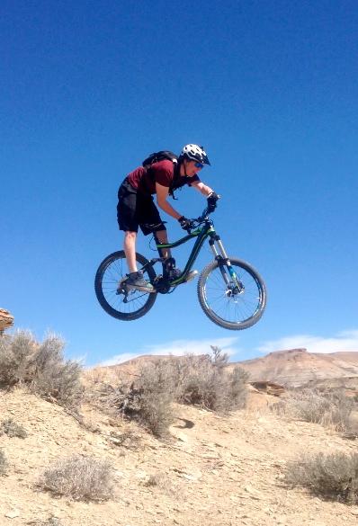 A mountain biker in mid-air performing a jump over rocky terrain against a clear blue sky. The rider is wearing a helmet and is dressed in shorts and a t-shirt, showcasing an adventurous outdoor activity. Sagebrush is visible in the foreground. Wilkins Peak Trails mountain bike trail.