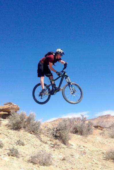 A cyclist performing a jump on a mountain bike over a rocky terrain, surrounded by sparse vegetation and a clear blue sky in the background. Wilkins Peak Trails mountain bike trail.