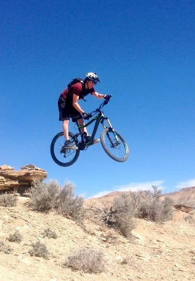 A mountain biker performing a jump over rocky terrain against a clear blue sky. The rider is wearing a helmet and athletic gear, showcasing action and athleticism as they soar above the ground with their bike. Surrounding the area are sparse shrubs and desert-like landscape. Wilkins Peak Trails mountain bike trail.