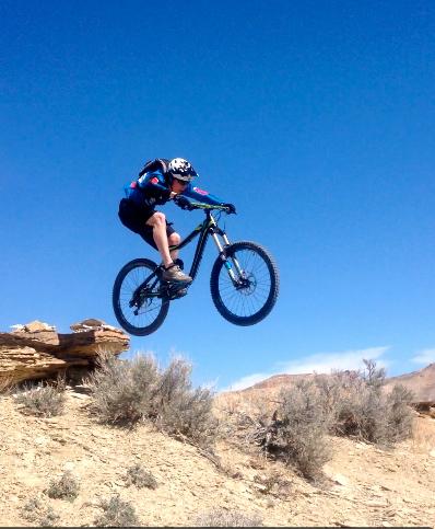 A person in a blue and black cycling outfit is mid-air, jumping off a rocky ledge on a mountain bike. The background features clear blue skies and sparse desert vegetation, highlighting an outdoor biking adventure. Wilkins Peak Trails mountain bike trail.
