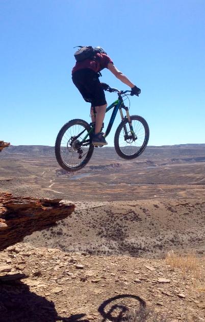 A mountain biker performing a jump off a rocky ledge, with a vast desert landscape visible in the background under a clear blue sky. Wilkins Peak Trails mountain bike trail.
