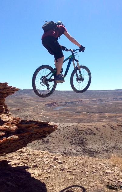 A mountain biker in mid-air, performing a jump off a rocky ledge with a vast landscape of hills and valleys in the background under a clear blue sky. Wilkins Peak Trails mountain bike trail.
