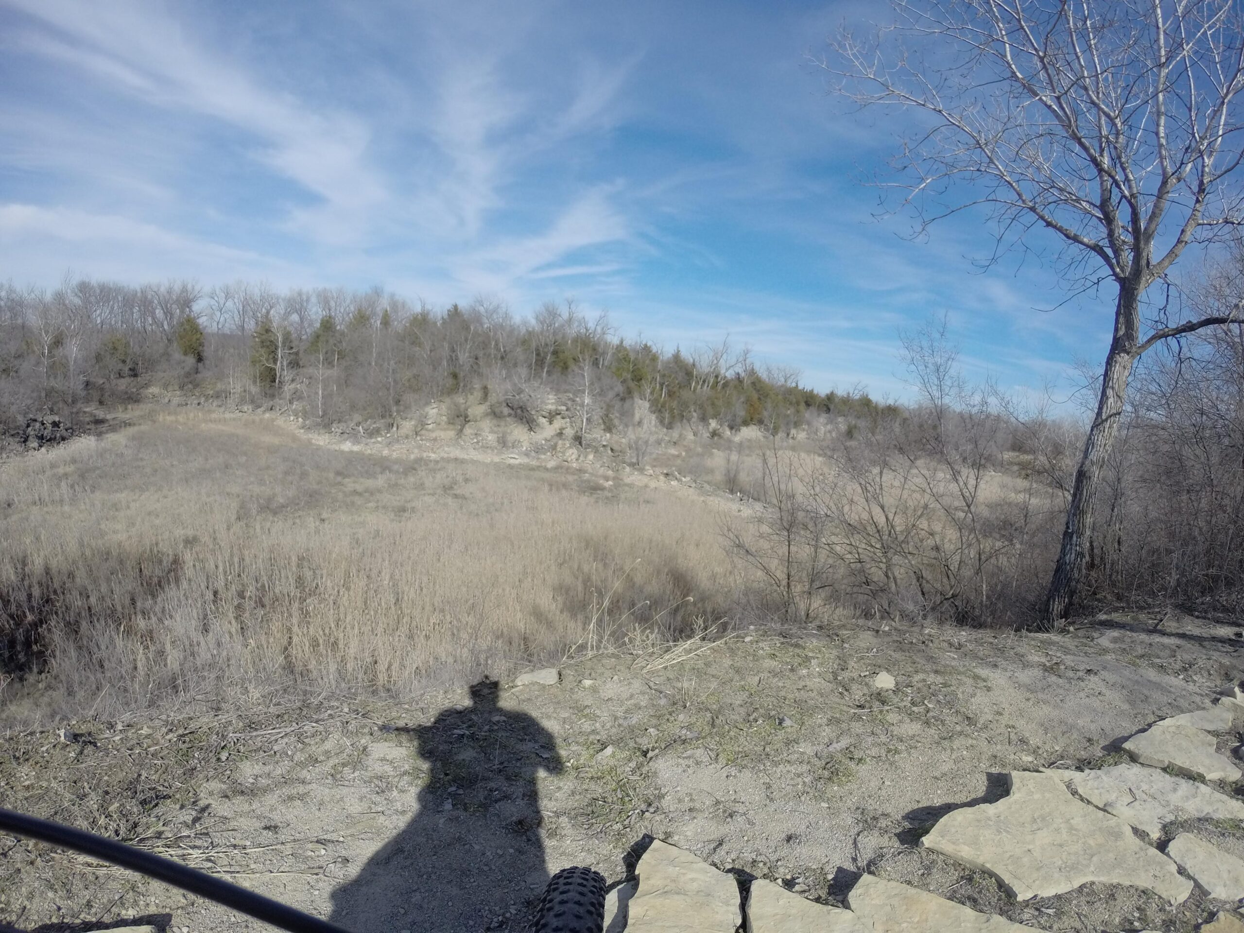A landscape view featuring a rocky terrain with dry grasses and sparse trees, under a clear blue sky with wispy clouds. The foreground includes a shadow of a person and stone pathways leading to the nature scene. Melvern Riverfront Trails mountain bike trail.