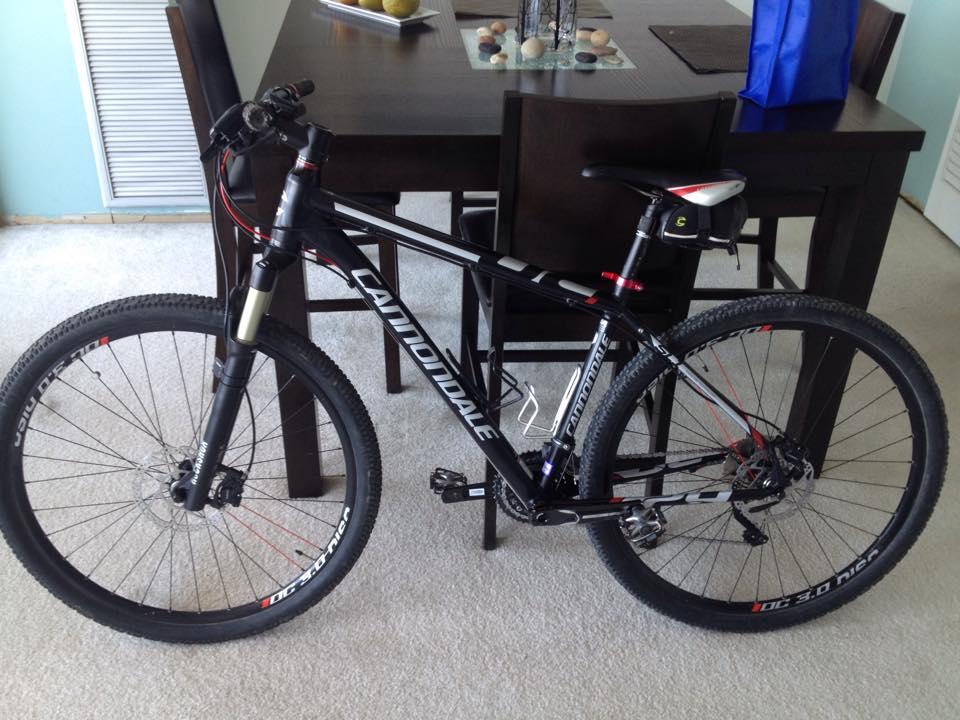 Cannondale Trail SL 29 1: A black Cannondale mountain bike is leaning against a dining table. The bike features thick tires and a sleek frame, with visible brand markings. The background includes a light-colored carpet, a dining table with decorative items, and a blue bag.
