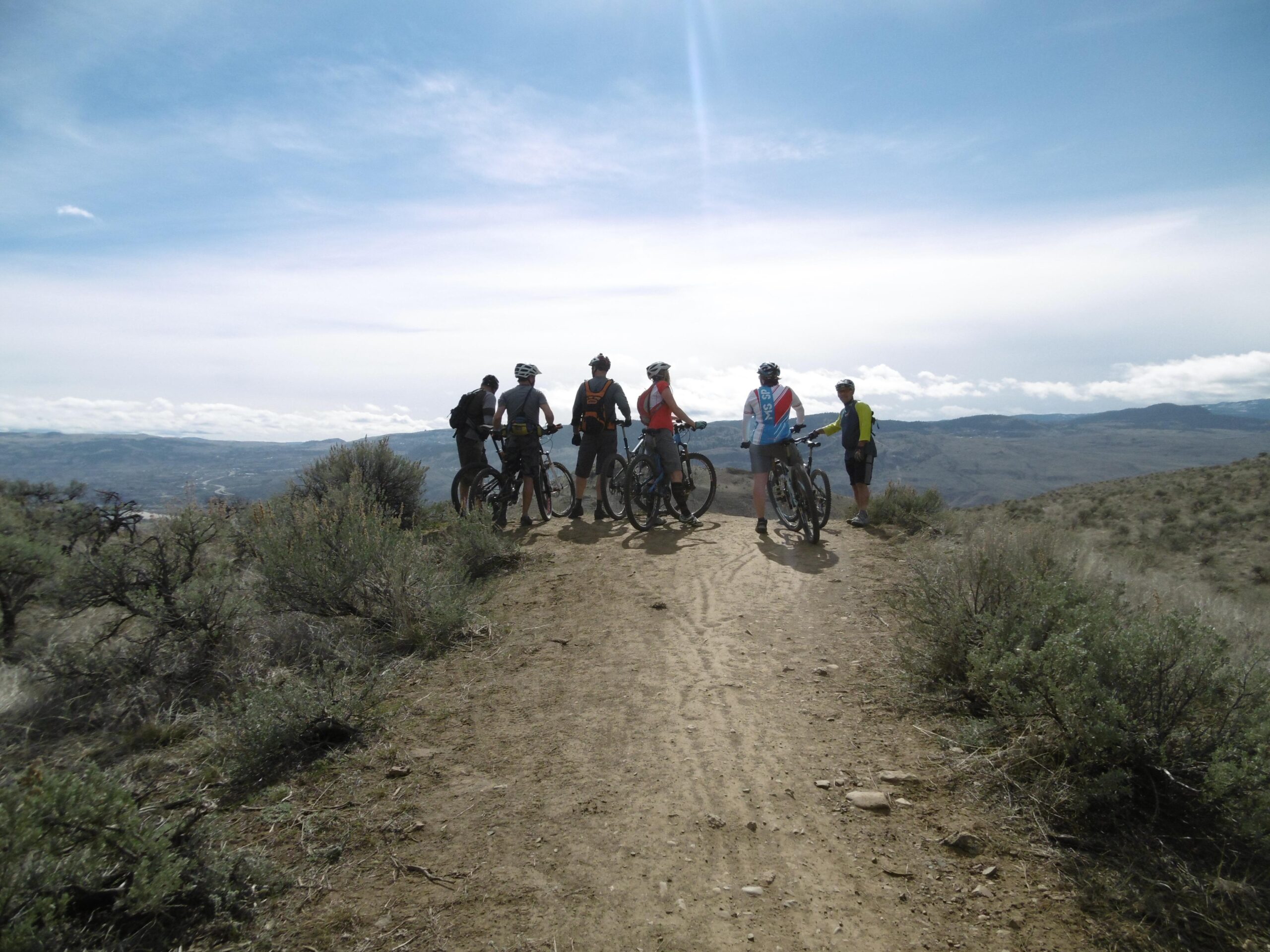 A group of five mountain bikers pause on a dirt trail surrounded by sparse vegetation, overlooking a scenic landscape with mountains and clouds in the background. The riders are dressed in outdoor cycling gear and are positioned with their bikes, enjoying a moment of rest while taking in the view. Batchelor Heights mountain bike trail.