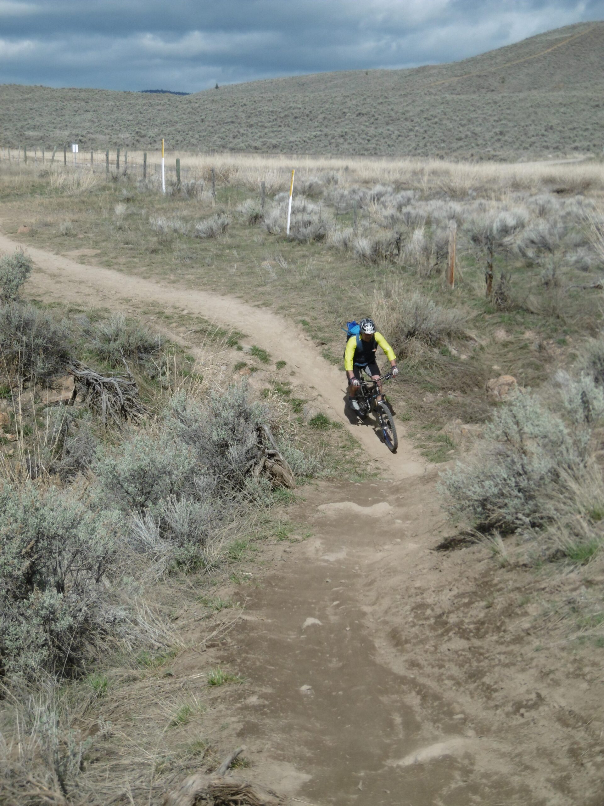 A mountain biker in a bright yellow long-sleeve shirt navigates a dirt trail surrounded by sagebrush, with rolling hills and a cloudy sky in the background. Batchelor Heights mountain bike trail.
