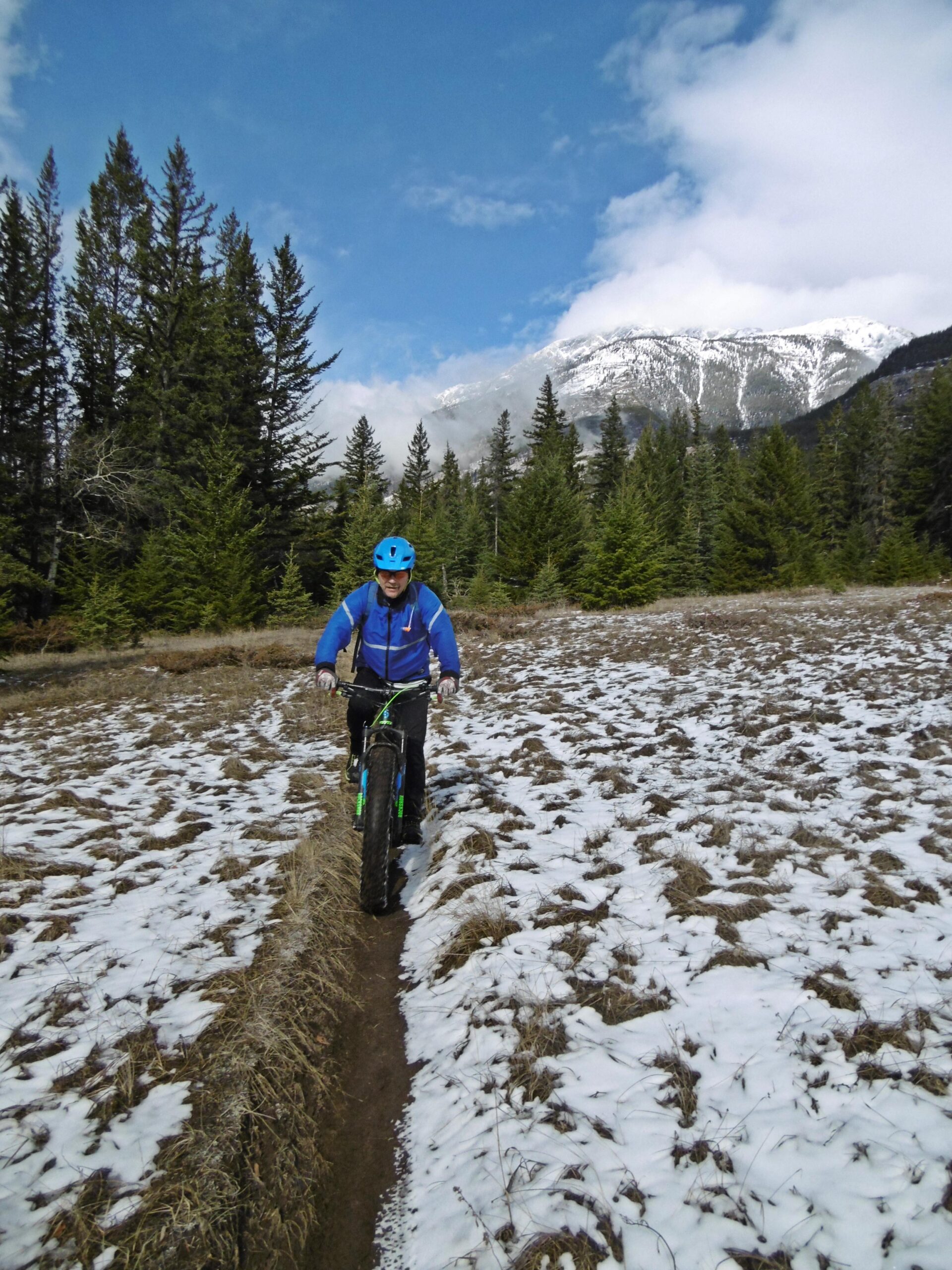 A person riding a fat bike on a snowy trail, surrounded by tall evergreen trees and mountains in the background. The sky is partly cloudy, with patches of blue visible. The cyclist is wearing a blue jacket and a blue helmet. The Overlander mountain bike trail.