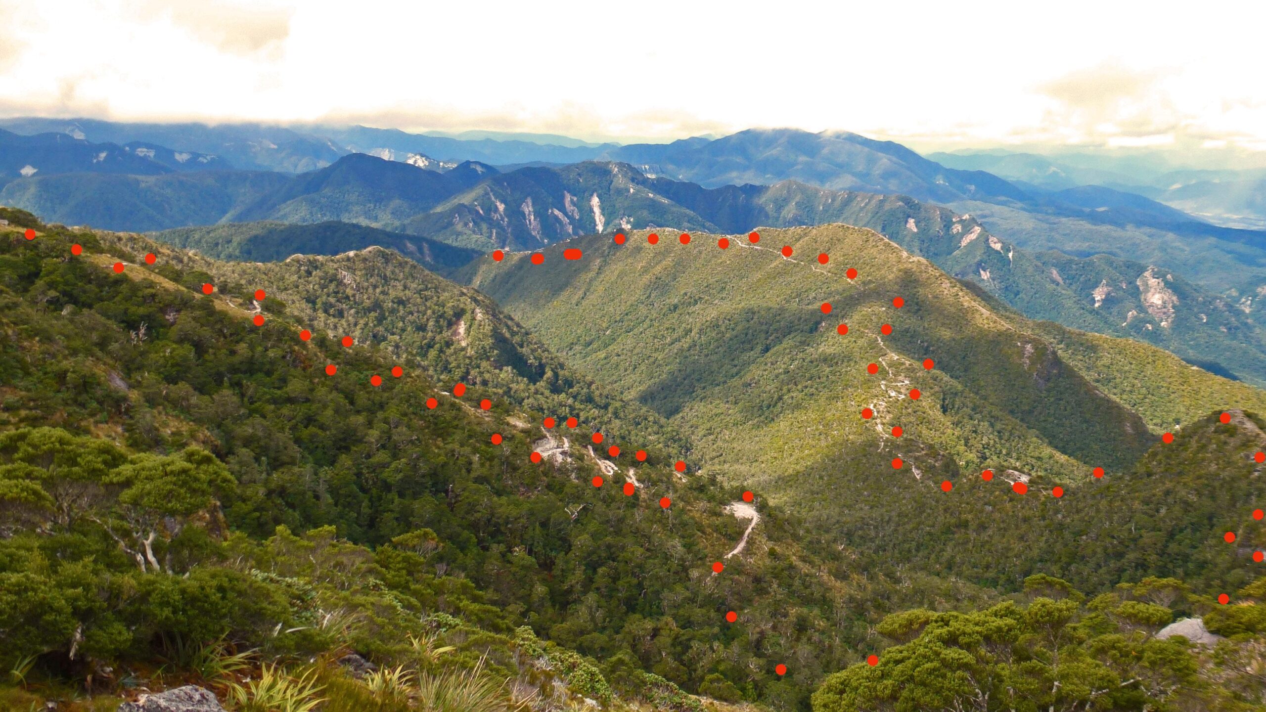 A panoramic view of lush mountainous terrain, featuring rolling hills covered in dense green foliage. A winding trail is visible, marked by a series of red dots along its path. The background showcases a range of mountains under a partly cloudy sky, adding depth to the serene landscape. The Old Ghost Road mountain bike trail.