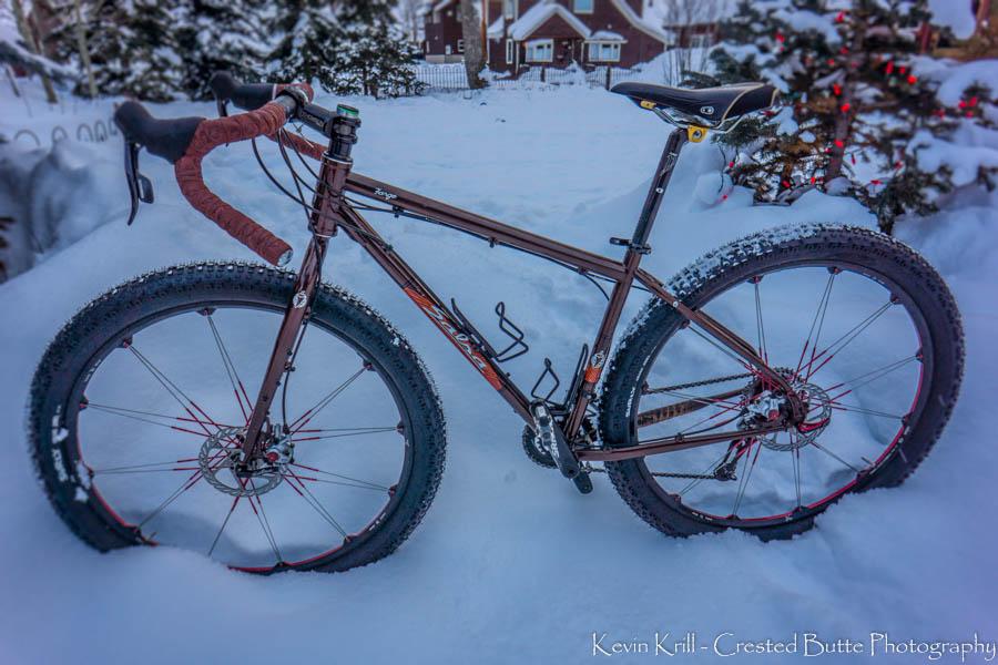Salsa Fargo: A brown fat bike with wide tires positioned on a snowy landscape, featuring a simple design and black and red accents. In the background, there are hints of a residential area with snow-covered trees and holiday decorations.
