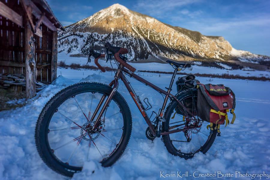 Salsa Fargo: A fat bike resting on snowy ground with a rugged mountain in the background, bathed in sunlight. The bike features wide tires and is equipped with a red and yellow pannier bag. An old wooden structure is partially visible to the left, complementing the winter landscape.