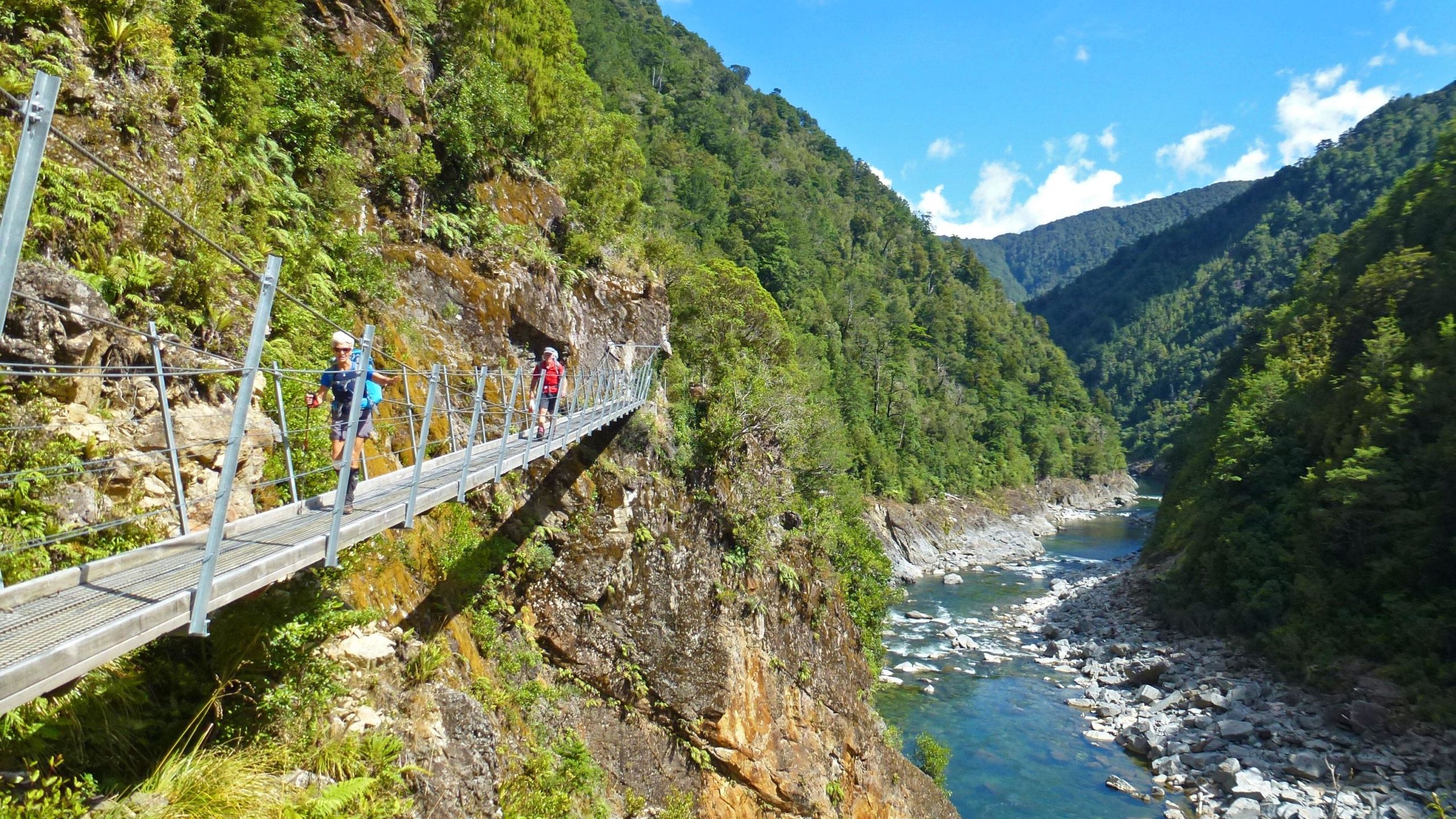 A suspension bridge traversing a deep valley, surrounded by lush green vegetation and steep mountains. Two hikers, one wearing a blue shirt and the other in red, are crossing the bridge, while a flowing river is visible below among the rocky banks. The sky above is clear with some clouds. The Old Ghost Road mountain bike trail.
