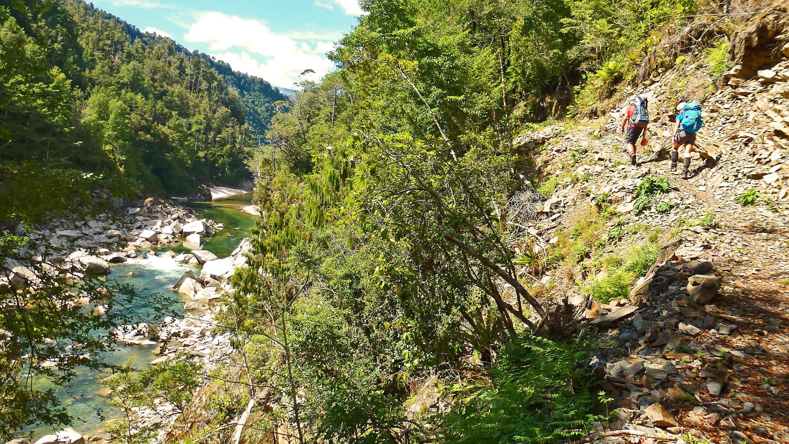 Two hikers navigate a rocky trail along a riverbank, surrounded by lush green forests and mountains under a bright blue sky. The Old Ghost Road mountain bike trail.