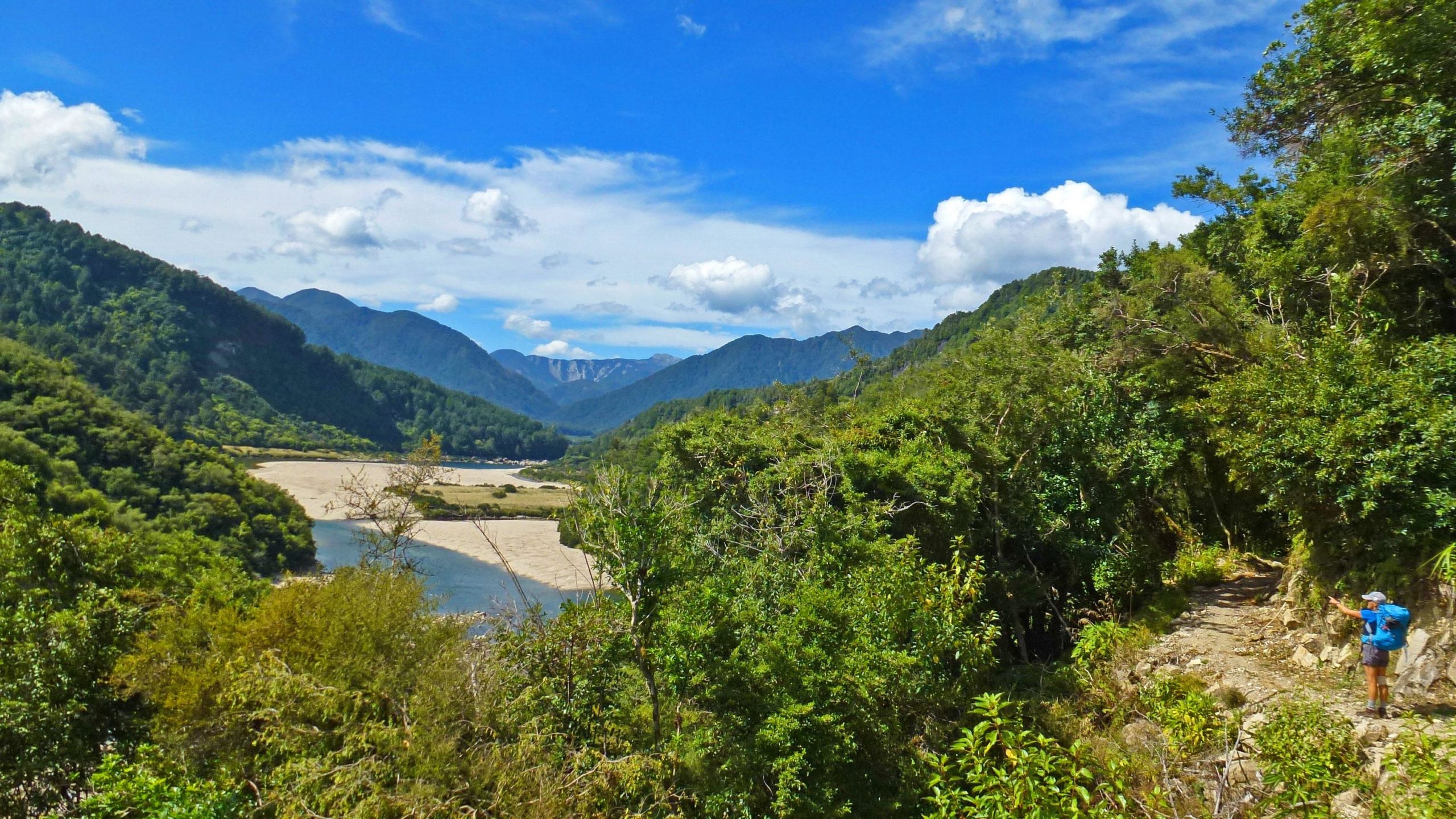 A scenic landscape featuring lush green hills and mountains under a bright blue sky with white clouds. A river winds through the valley, which is dotted with sandy areas. A hiker, dressed in a blue shirt and shorts, stands on a rocky path, gesturing towards the view, surrounded by dense foliage. The Old Ghost Road mountain bike trail.