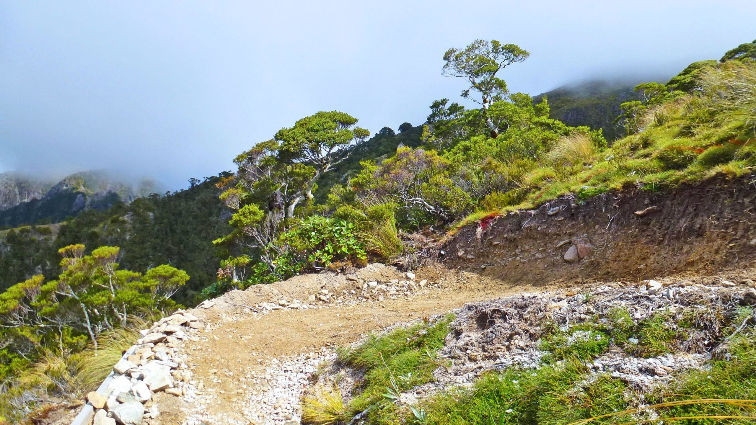 A winding dirt path curves along a hillside, surrounded by lush greenery and trees. The background features a misty landscape with distant mountains partially obscured by clouds. The terrain shows a mix of bare earth and small rocks, indicating natural scenery typical of a mountainous area. The Old Ghost Road mountain bike trail.