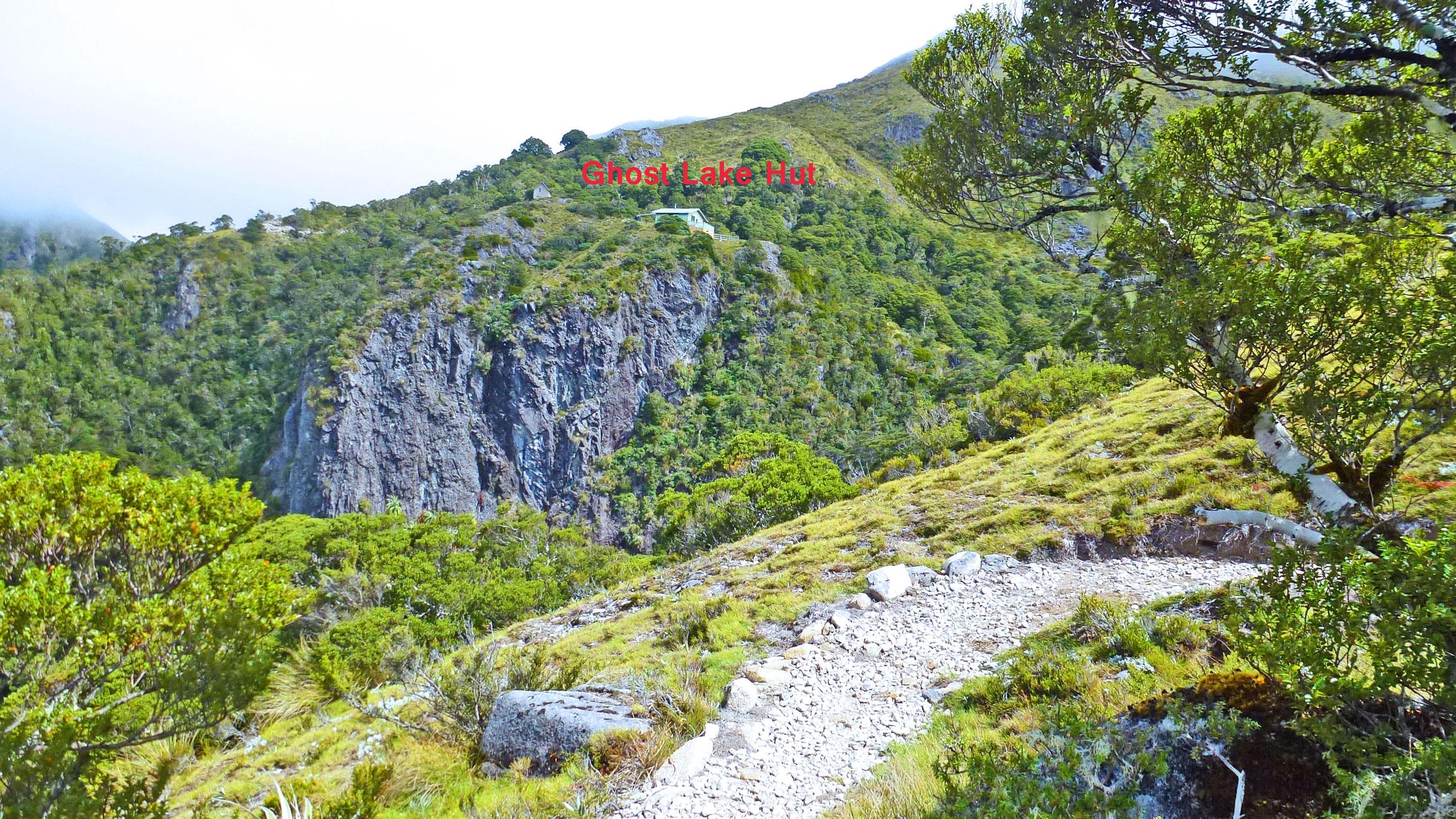 Scenic view of a lush green hillside featuring Ghost Lake Hut prominently labeled in red. The landscape includes rocky cliffs, dense foliage, and a winding gravel path leading through the foreground, capturing the natural beauty of the area. The Old Ghost Road mountain bike trail.