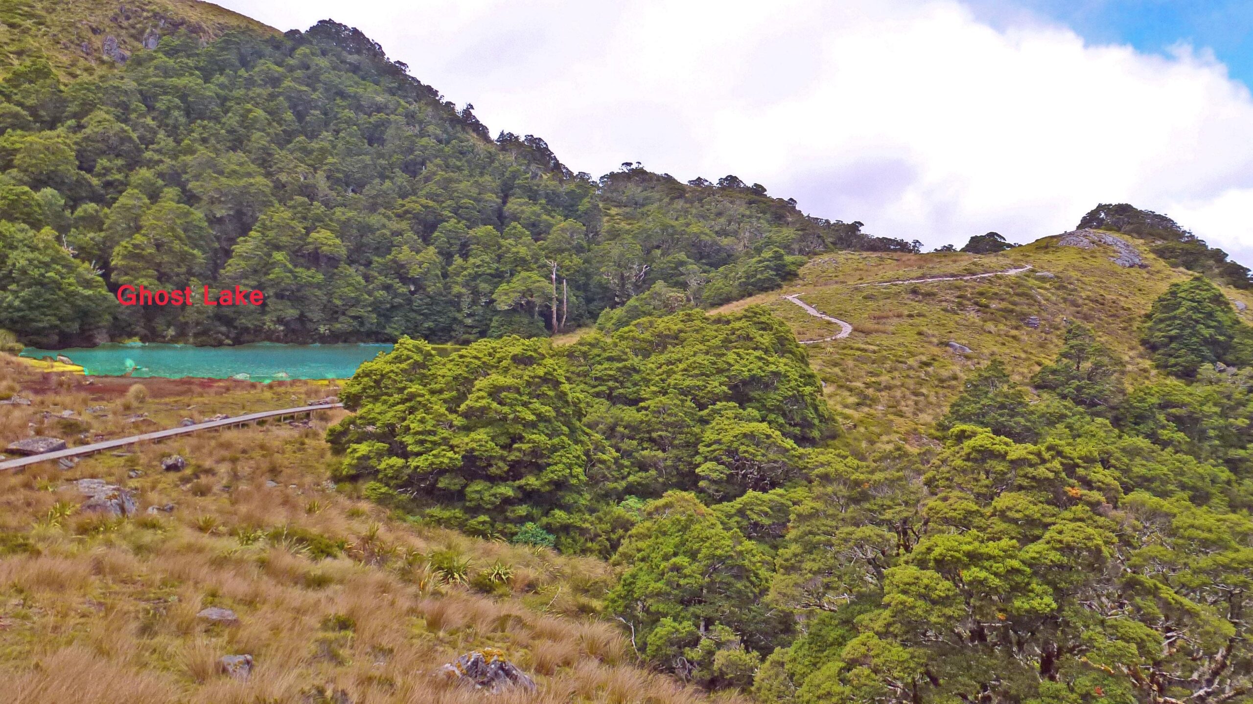 Scenic view of Ghost Lake surrounded by lush greenery and hills, with a pathway winding through the landscape. The lake features a distinct turquoise color, contrasting against the earthy tones of the grass and rocks. The Old Ghost Road mountain bike trail.