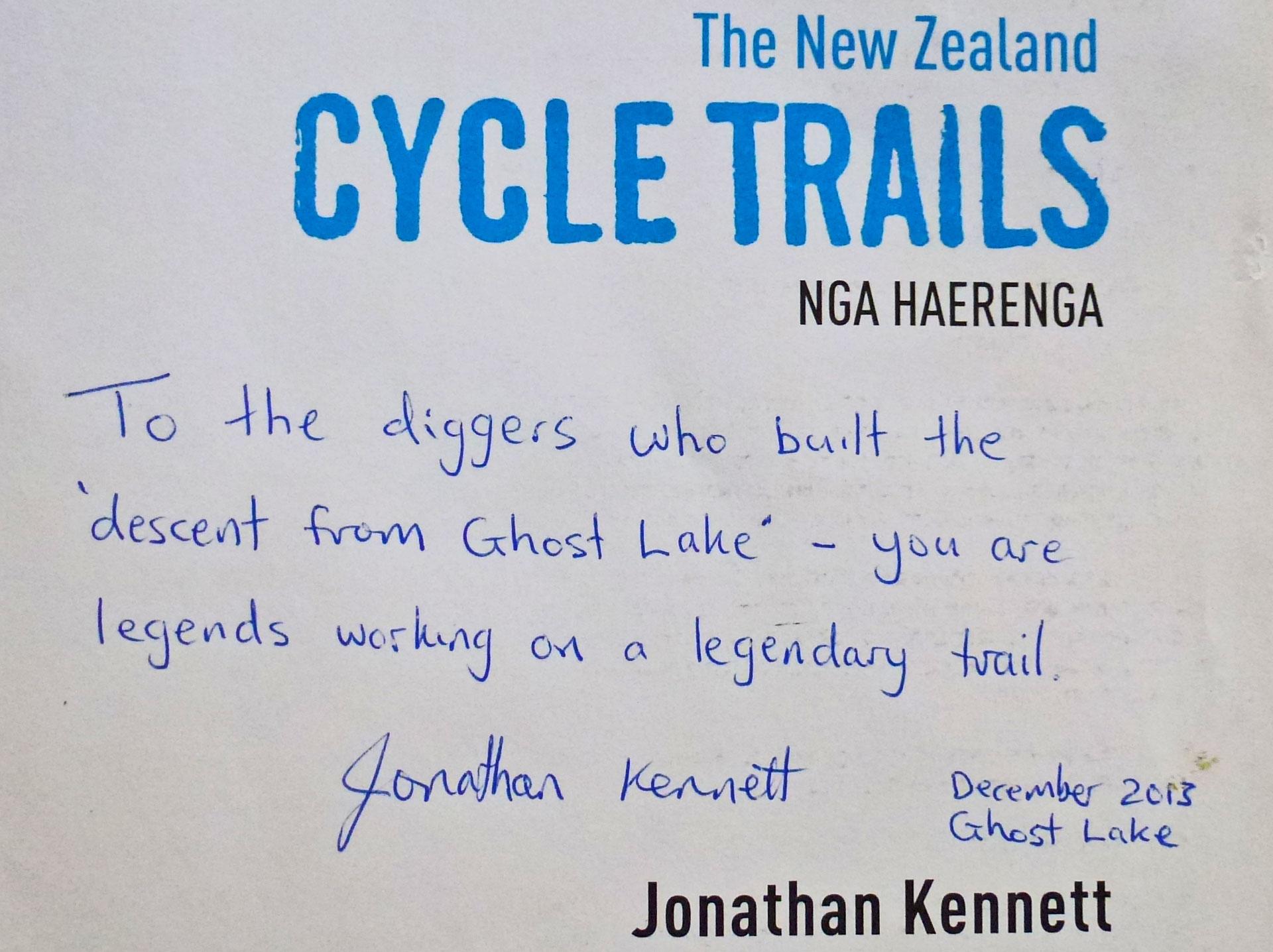 A handwritten note thanking the diggers who built the "descent from Ghost Lake," recognizing their contributions to a legendary trail. The note is signed by Jonathan Kennett and dated December 2013. The image also features the title "The New Zealand Cycle Trails" at the top. The Old Ghost Road mountain bike trail.