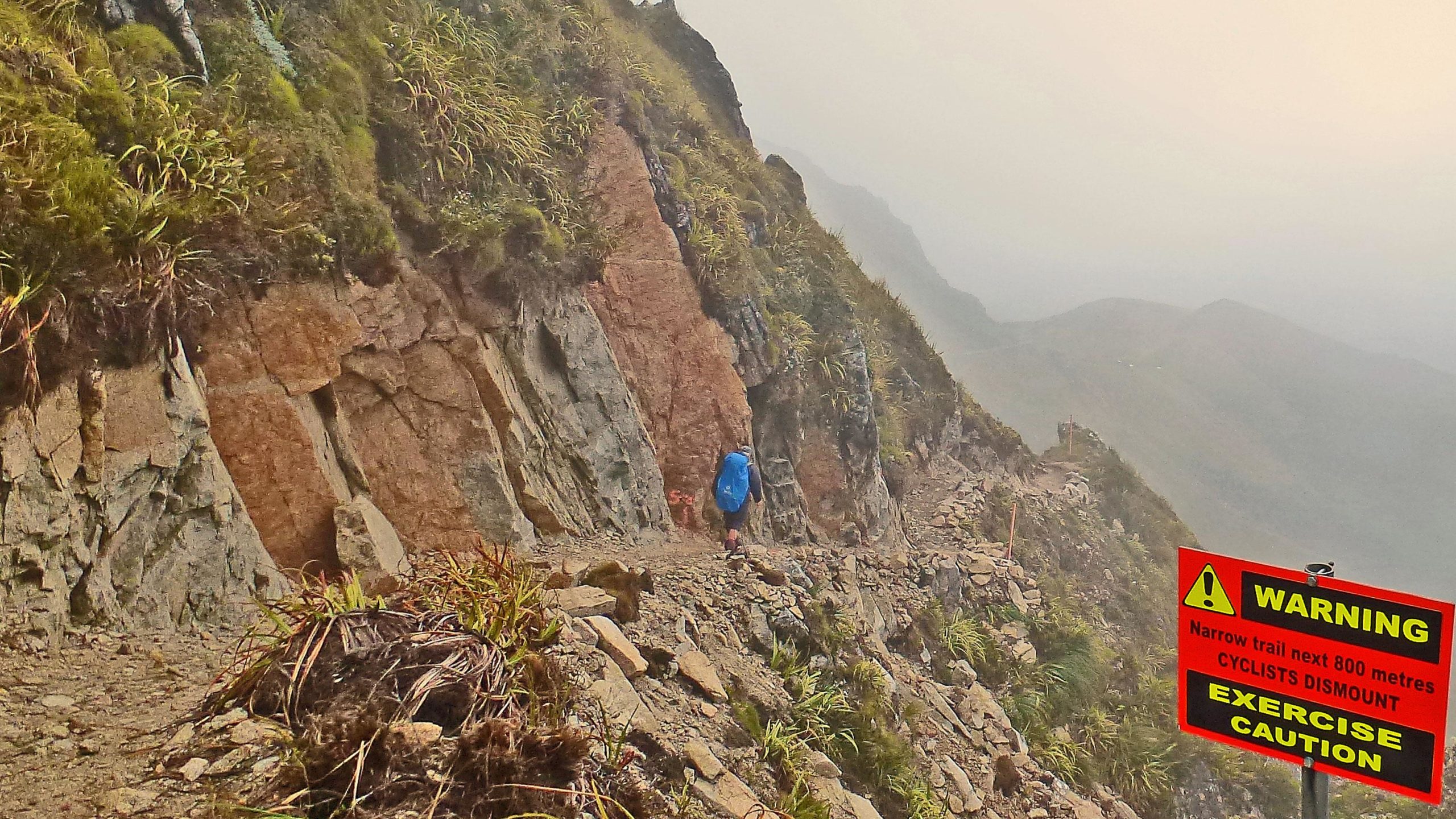 A hiker with a blue backpack traverses a narrow mountain trail lined by rocky cliffs and greenery, with fog obscuring the distant landscape. A bright warning sign indicates that the trail is narrow for the next 800 meters and advises cyclists to dismount and exercise caution. The Old Ghost Road mountain bike trail.