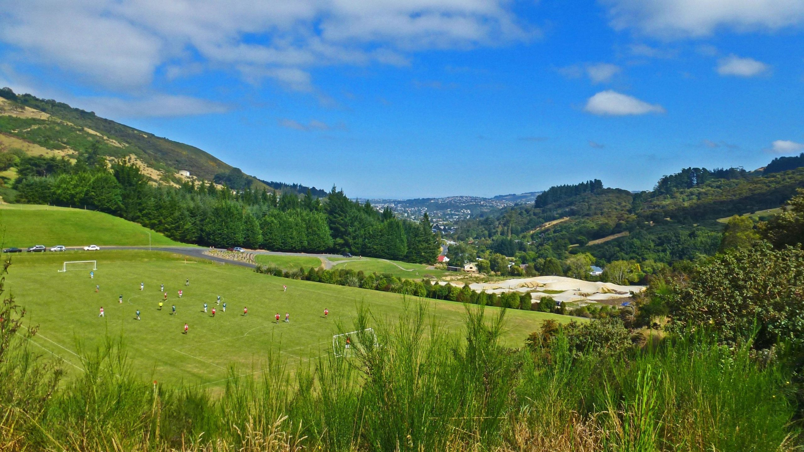 A panoramic view of a green soccer field filled with players, set against a backdrop of rolling hills and blue sky. Lush trees border the field, and a distant cityscape can be seen on the horizon. Bethunes Gully mountain bike trail.