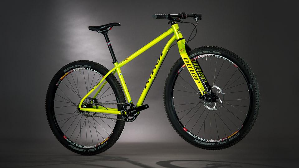 Niner S.I.R. 9: A bright yellow mountain bike with black wheels and frame components, posed against a dark background. The bike features a lightweight frame, suspension fork, and wide tires designed for off-road terrain.