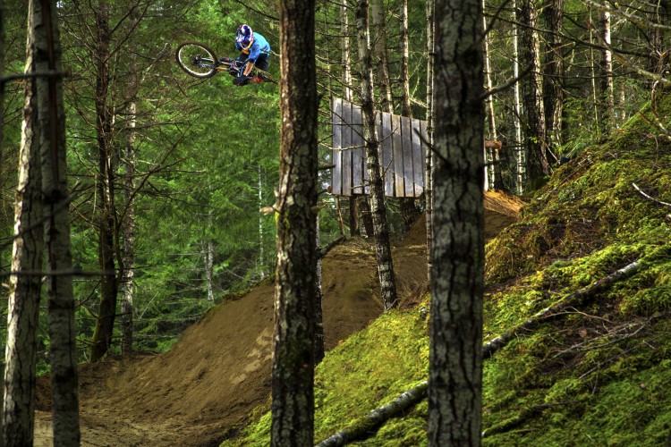 A mountain biker performing a trick in mid-air over a dirt ramp, surrounded by tall trees and dense greenery in a forest setting.