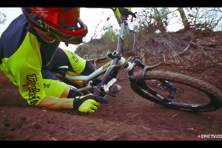 A mountain biker in a bright yellow and black outfit falls off their bike, leaning into the dirt while trying to regain control on a rugged trail surrounded by trees and underbrush.