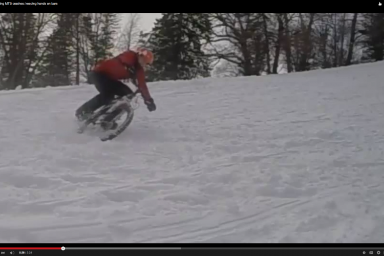 A person riding a mountain bike on snowy terrain, leaning into a turn while snow sprays up around the rear wheel. Trees are visible in the background, and the rider is wearing a red jacket and an orange helmet.