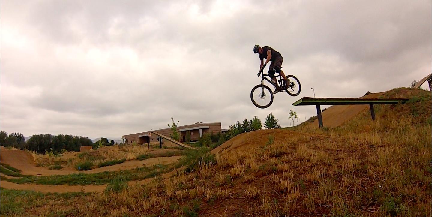 A cyclist performing a jump off a ramp at a dirt biking park, with a grassy area and cloudy sky in the background. Valmont Bike Park mountain bike trail.