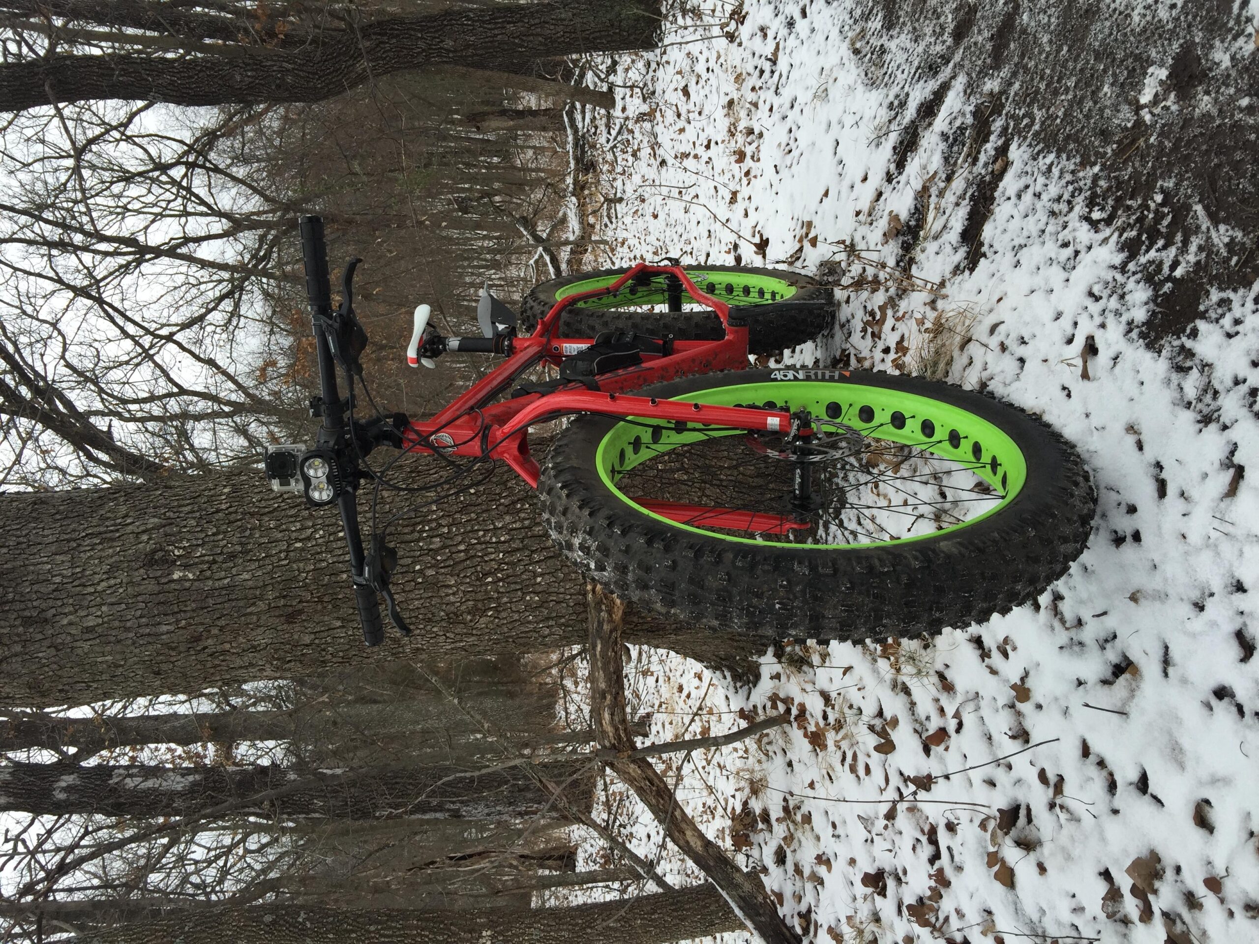 Salsa Mukluk: A fat tire bike with red and green accents parked on a snowy forest trail, surrounded by bare trees and fallen leaves.