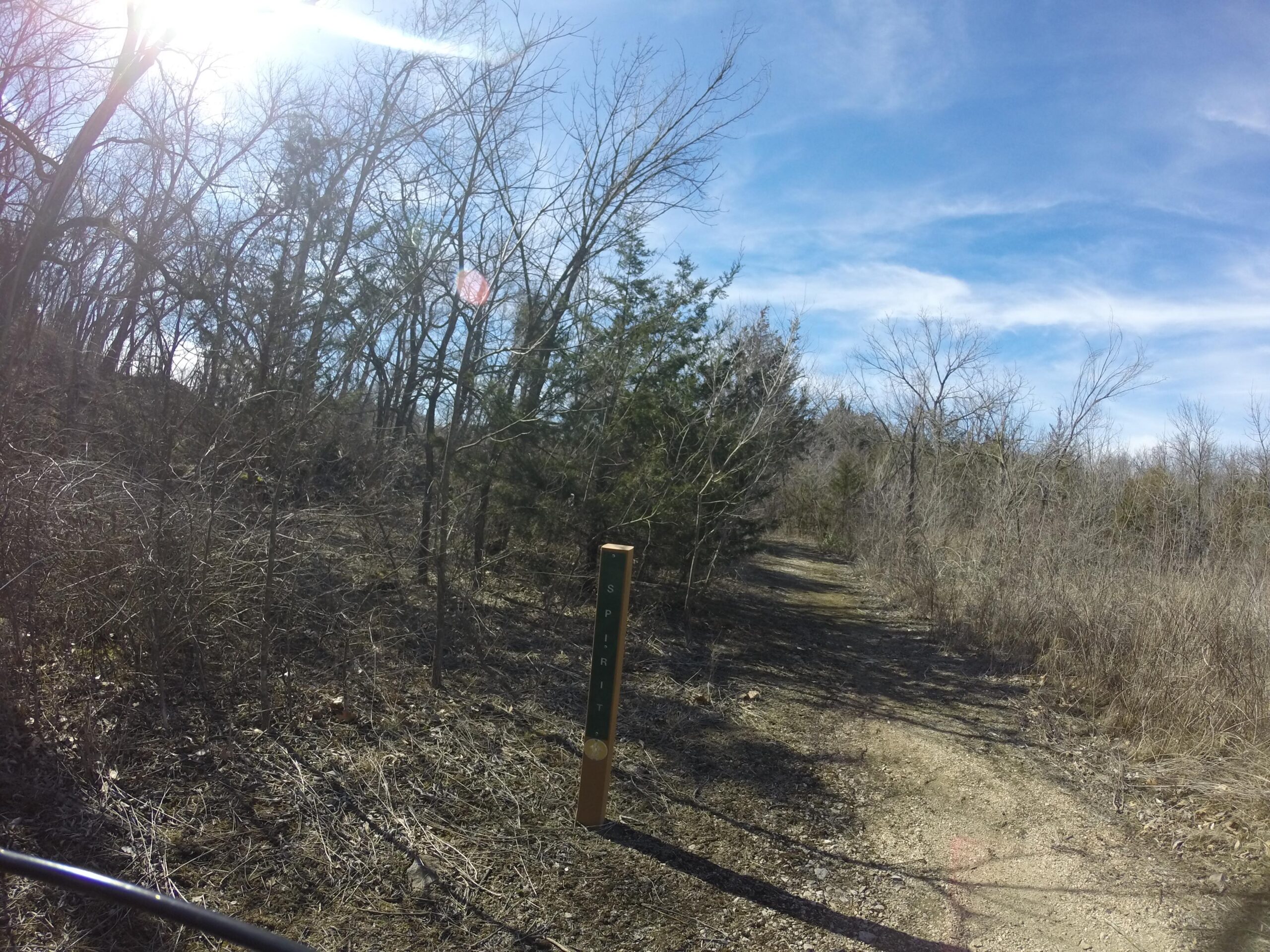 A dirt path meanders through a sparse wooded area, with tall, leafless trees on either side. A wooden trail sign stands on the left, indicating the "Spirit" trail. The sky above is bright blue with a few wispy clouds, and sunlight is shining down, creating a warm atmosphere. Melvern Riverfront Trails mountain bike trail.