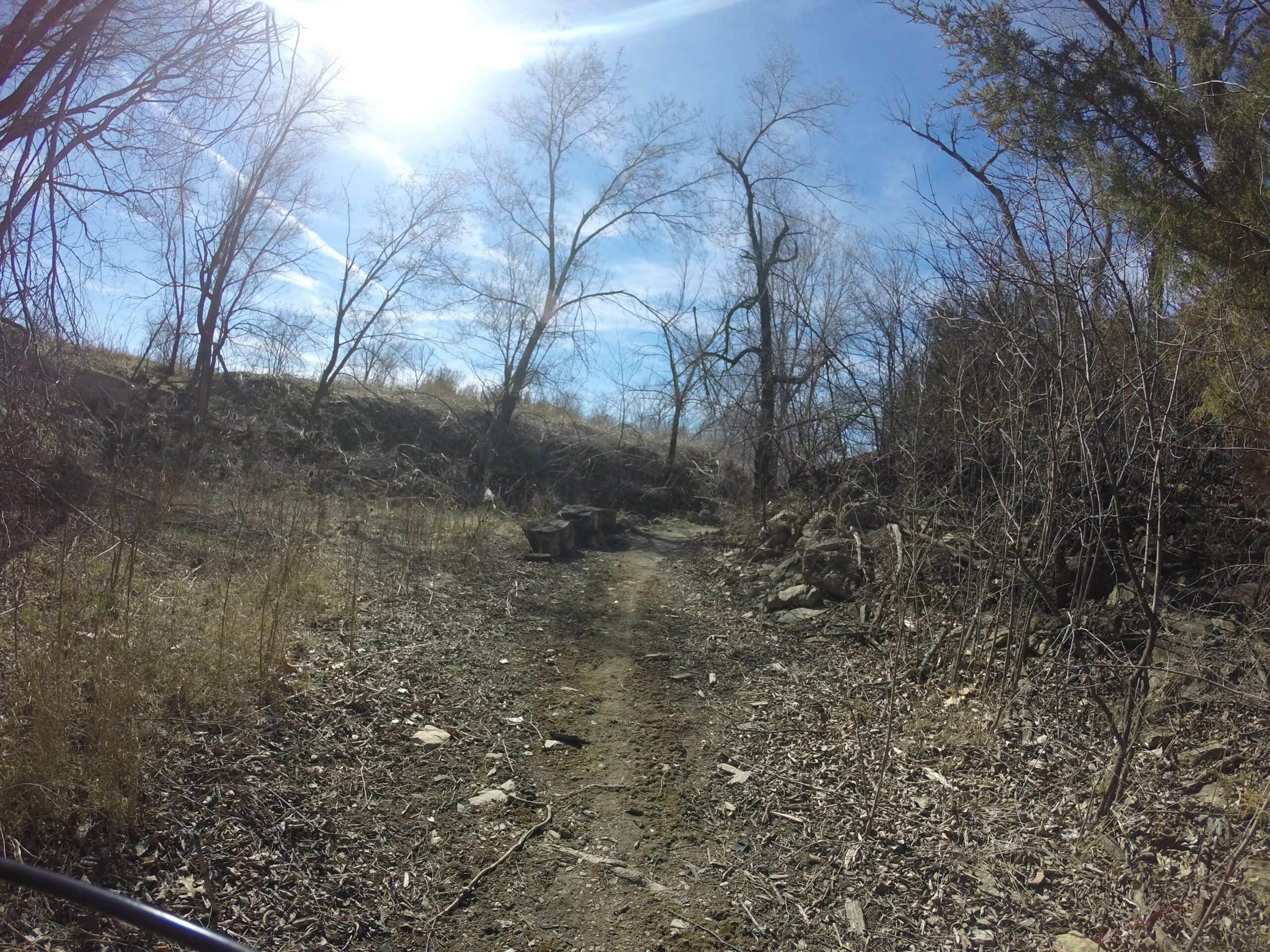 A dirt path winding through a sparse landscape with bare trees under a bright blue sky. The sun shines in the background, casting light on the dry ground and scattered rocks. Melvern Riverfront Trails mountain bike trail.