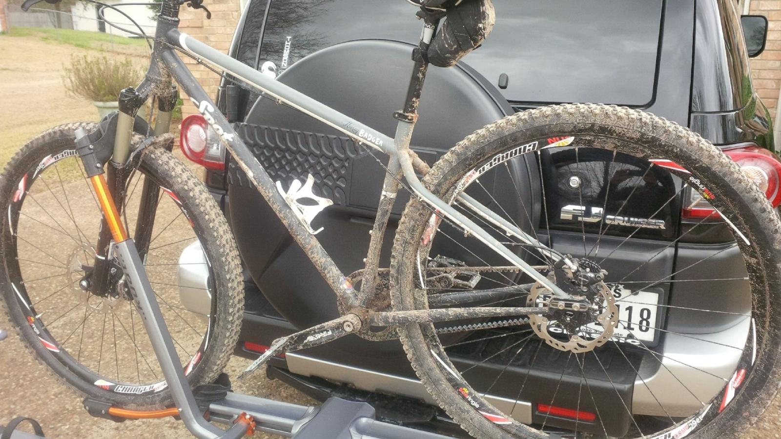 SPOT Honey Badger Singlespeed: A muddy mountain bike mounted on a car rack, with the vehicle's rear visible in the background. The bike features a gray frame, orange accents, and two heavily treaded wheels, indicating recent use in off-road conditions. Brown dirt and mud are caked on various parts of the bike, emphasizing its adventurous journey.