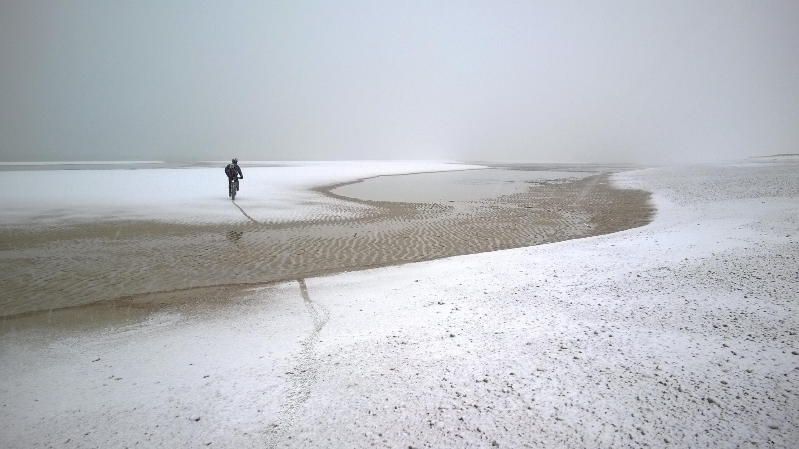 A person biking along a misty shoreline, with a landscape composed of wet sand and patches of snow. The sky is overcast and the visibility is low, creating a serene and quiet atmosphere. Sandy Neck Beach mountain bike trail.