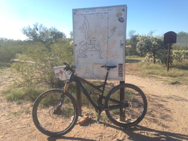 A mountain bike resting beside a trail map at a desert trailhead, with cacti and scrub vegetation in the background under a clear blue sky. The map displays various bike trails labeled "Fantasy Island." Fantasy Island mountain bike trail.