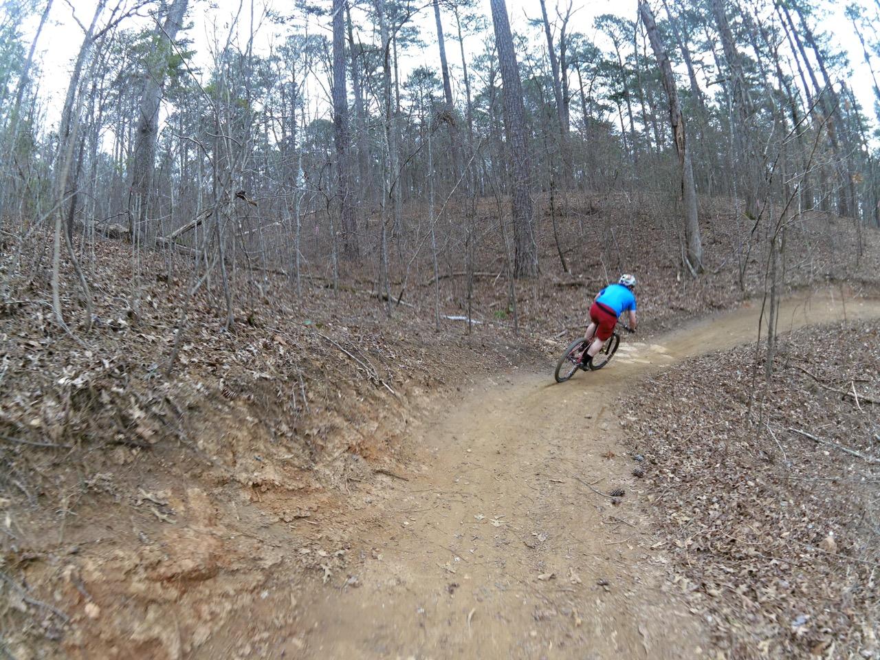 A cyclist wearing a blue shirt and red shorts navigates a winding dirt trail through a forested area. The path is surrounded by trees with sparse foliage, and fallen leaves cover the ground. The Mill mountain bike trail.