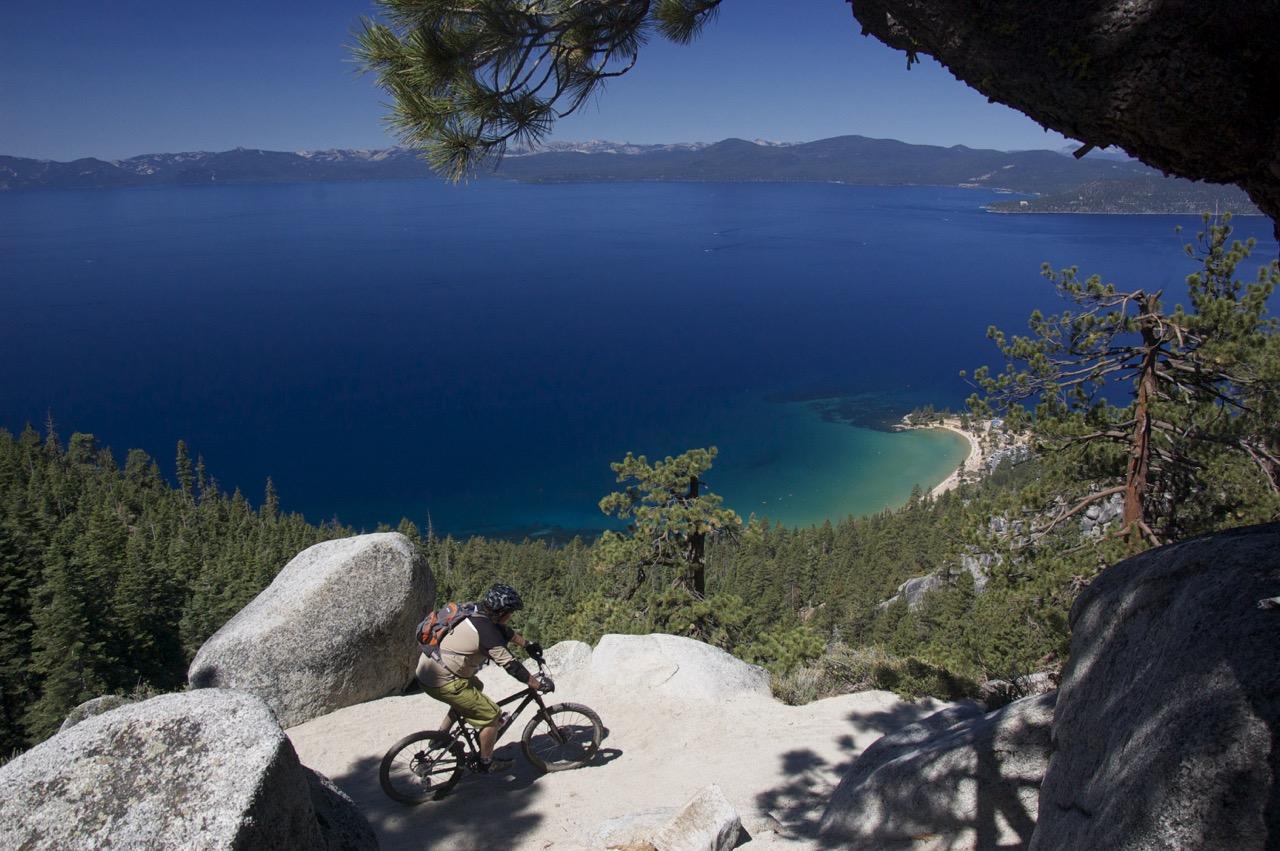 A mountain biker navigates a rocky trail overlooking a blue lake. The scene features dense green trees, boulders, and distant mountains under a clear sky. The calm water has shades of blue and green, with a sandy beach visible along the shore. Flume Trail mountain bike trail.
